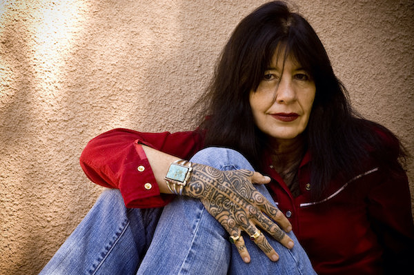 A photo of Joy Harjo wearing a red shirt and blue jeans.