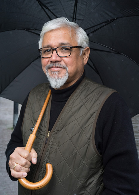 A photo of author Amitav Ghosh looking into the camera while holding an umbrella.