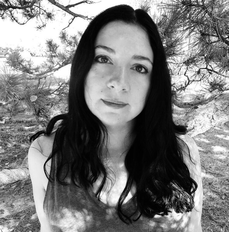 A photograph of author Alison Stine standing in front of a pine tree, looking into the camera.