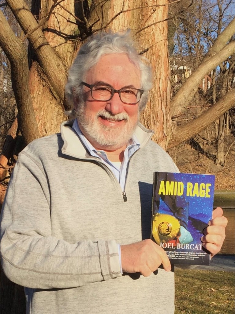 A photo of Joel Burcat standing in front of a tree, holding his novel Amid Rage.